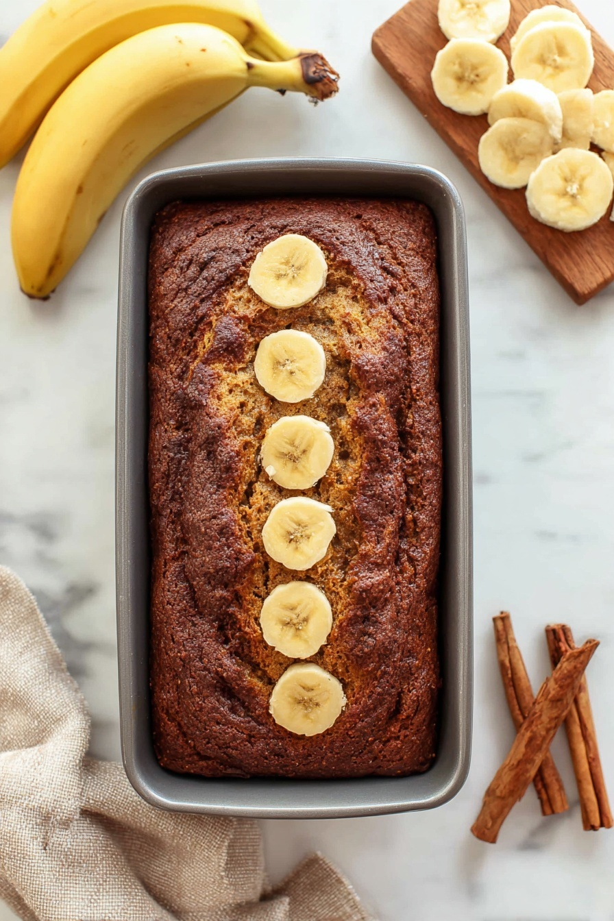 Cinnamon Swirl Banana Bread, moist banana bread with cinnamon, homemade banana bread with cinnamon swirl, easy cinnamon banana bread recipe, flavorful banana bread - A thick rectangular loaf of dark brown banana bread rests in a grey baking pan, with five round banana slices lined up in the center top of the bread. To the left, two whole yellow bananas lay curved on a white marbled surface. To the right, a wooden board holds several light yellow banana slices arranged in overlapping circles. At the bottom right corner, three brown cinnamon sticks rest on the white marbled surface, and a beige cloth is partially visible at the bottom left corner. photo taken with an iphone --ar 2:3 --v 7