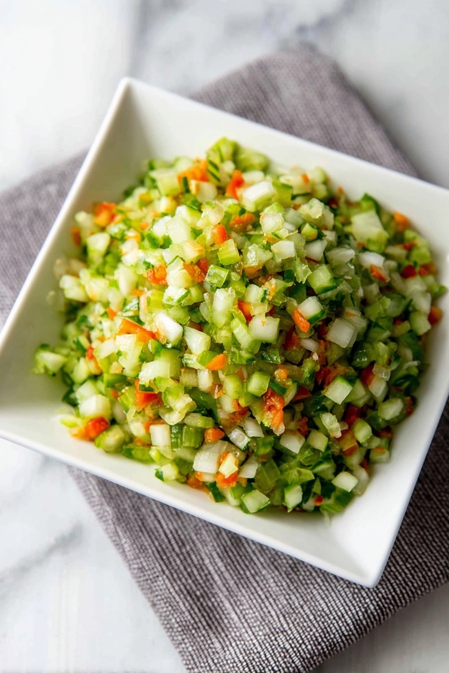 Dill Pickle Salsa, tangy salsa recipe, crunchy pickle salsa, fresh salsa with dill pickles, easy pickle salsa - A close-up view of finely chopped vegetables in a white square bowl placed on a grey cloth on a white marbled surface. The mix has small pieces with mostly green colors from celery or cucumber, bright orange bits from carrots, and some white pieces from onions, creating a fresh and colorful look. The texture appears crisp and moist, with the small vegetable pieces piled up in layers that give a sense of volume and freshness. photo taken with an iphone --ar 2:3 --v 7