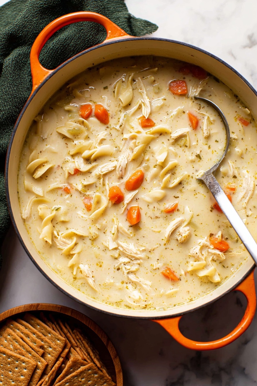 Creamy Chicken Noodle Soup, hearty chicken soup recipe, easy creamy chicken noodle soup, comforting chicken soup, best chicken noodle soup - A close-up of a white pot filled with creamy chicken noodle soup, showing a thick light beige broth mixed with small bright orange carrot slices, soft shredded white chicken pieces, and flat light yellowish noodles scattered throughout. A silver ladle is partially dipped in the soup, resting on the right side of the pot. The pot has an orange handle on each side and sits on a white marbled surface. Below the pot, there is a stack of brown crackers arranged neatly in a small wooden bowl. A dark green cloth is folded behind the pot, adding contrast to the scene. Photo taken with an iphone --ar 2:3 --v 7