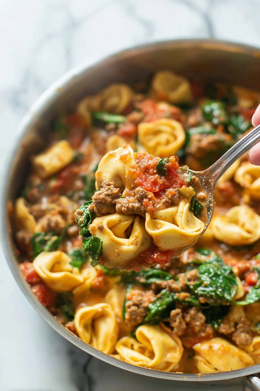 Creamy Sausage Tortellini Skillet, sausage tortellini dinner, easy one-pan pasta recipe, hearty sausage skillet, quick creamy pasta - A close-up image of a cooked pasta dish in a silver pan shows three main layers. The bottom layer is pasta tortellini, small ring-shaped with light yellow color and slightly ruffled edges. The middle layer features ground meat chunks mixed with pieces of red tomato, all coated in a creamy, light brown sauce. The top layer has fresh green spinach leaves scattered throughout, adding vibrant color and texture. A woman's hand holding a silver spoon lifts a portion of the pasta, meat, tomato, and spinach mixture above the pan. The background is a white marbled surface. photo taken with an iphone --ar 2:3 --v 7