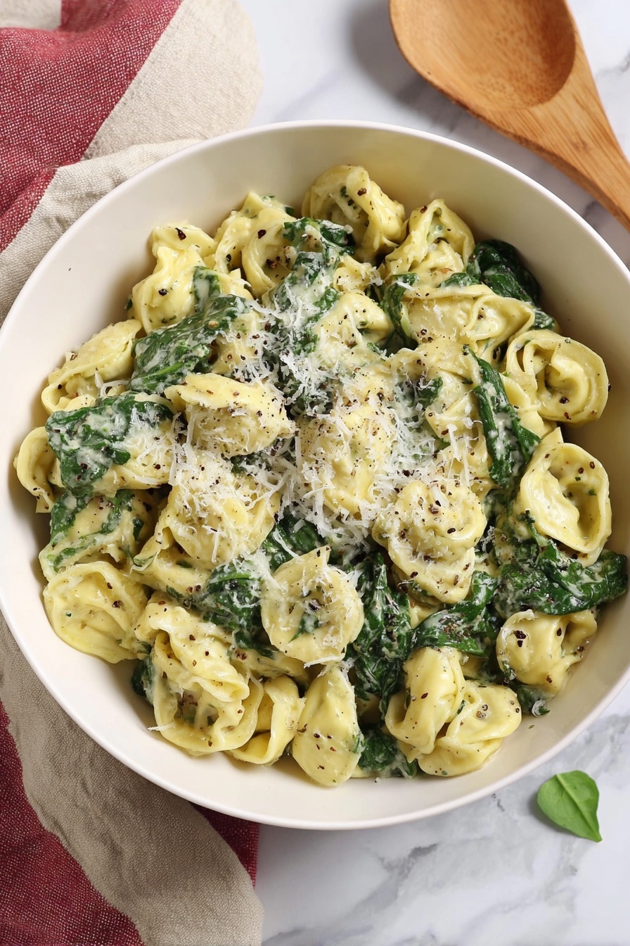Cheese Tortellini with Pesto and Spinach, quick pasta recipes, easy vegetarian dinner, healthy tortellini ideas, Italian pasta with pesto - A white bowl filled with three layers of tortellini pasta that is light yellow, mixed with dark green spinach leaves, and coated in a creamy sauce with flecks of black pepper and herbs. On top, there is a light sprinkle of grated white cheese finely covering some pasta and spinach. The bowl sits on a white marbled surface with a beige and red cloth folded beside it, and a wooden spoon rests nearby, all softly lit in natural light. Photo taken with an iphone --ar 2:3 --v 7