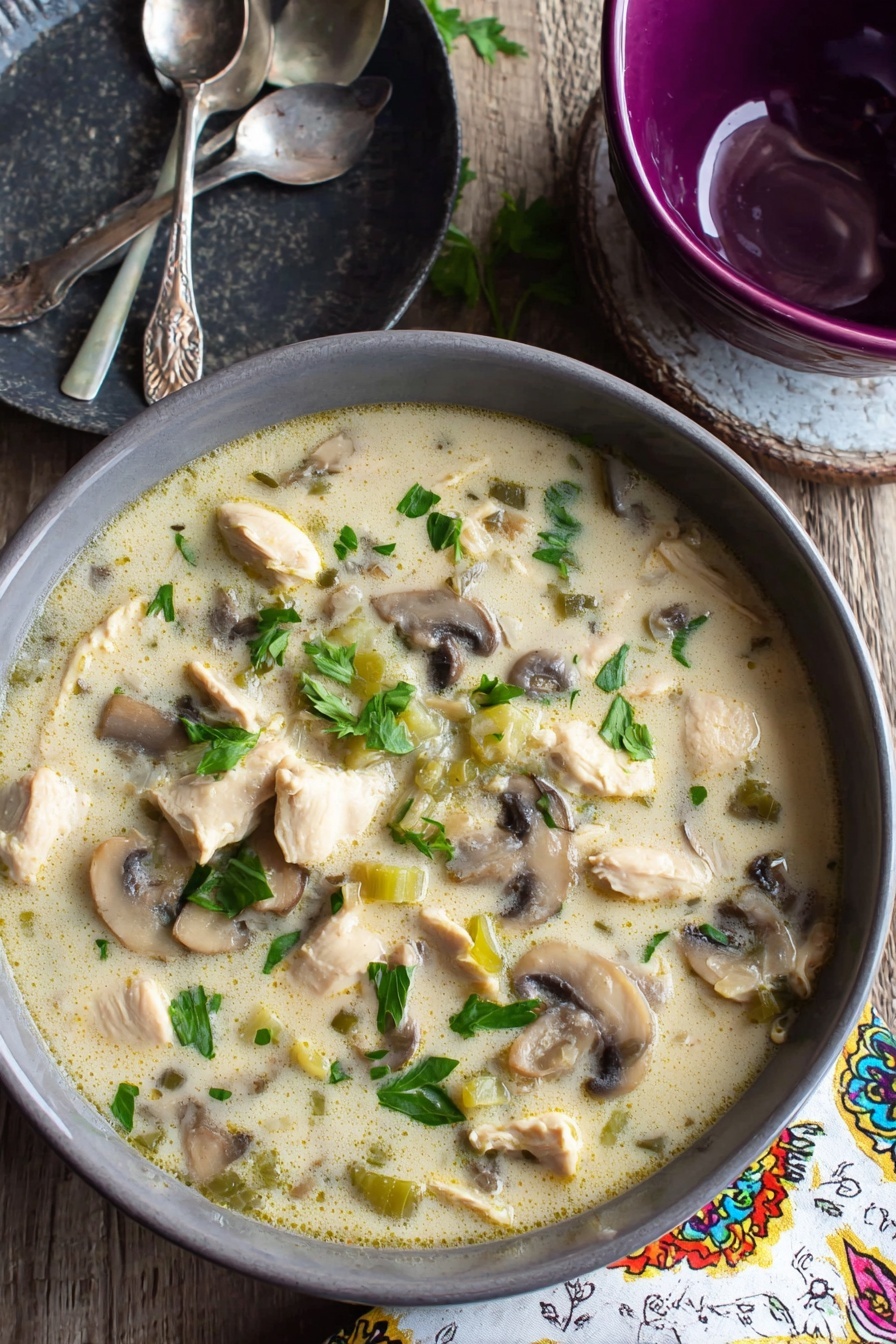 Creamy Chicken Mushroom Soup, hearty mushroom chicken soup, comforting chicken and mushroom dinner, easy creamy chicken mushroom recipe, cozy mushroom chicken bowl - A gray bowl filled with creamy soup that has chunks of light beige cooked chicken, sliced brown and white mushrooms, and small pieces of green celery, all mixed in a pale, creamy broth with green parsley leaves scattered on top, sitting on a wooden table next to a dark gray plate with two silver spoons and a purple bowl, with a white napkin that has colorful patterns to the side, the surface beneath is a white marbled texture photo taken with an iphone --ar 2:3 --v 7