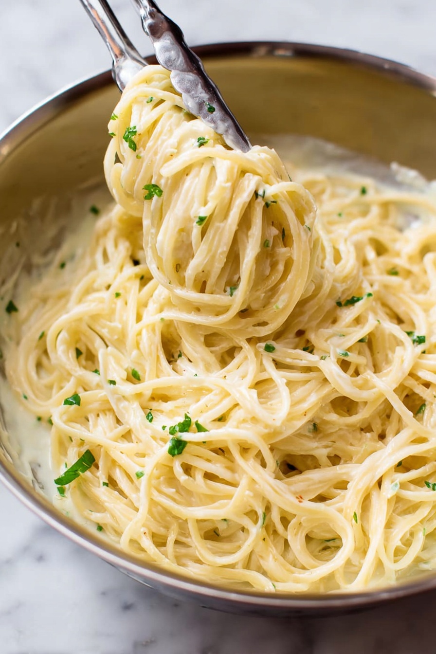 Garlic Parmesan Pasta in One Pot, easy one pot pasta, creamy garlic pasta, quick weeknight dinner, cheesy pasta recipe - A close-up view of a stainless steel bowl filled with creamy spaghetti pasta covered in a smooth, pale yellow sauce. The pasta strands are thick and smoothly coated, twirled around a pair of metal tongs that are lifting some of the noodles from the bowl. Small bits of green herbs are sprinkled lightly over the pasta, adding a touch of color contrast. The background is a white marbled texture. photo taken with an iphone --ar 2:3 --v 7