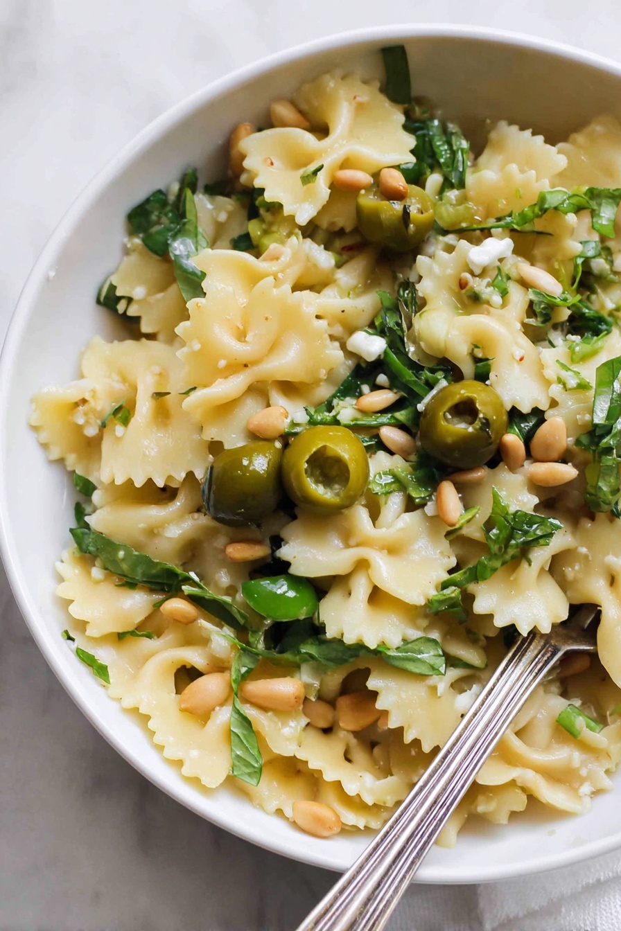 Dirty Martini Pasta Salad, savory pasta salad with olives and herbs, easy pasta salad recipes, briny pasta salad ideas, crowd-pleasing pasta salad - A close-up view of a white bowl filled with one layer of wavy-edged pasta pieces that are light yellow in color. Scattered on top are halved green olives, fresh green leafy herbs, and a few light brown pine nuts. There are also small pieces of a creamy white ingredient and some garlic cloves. A silver fork rests inside the bowl at the bottom right corner. The bowl is placed on a white marbled surface. Photo taken with an iphone --ar 2:3 --v 7