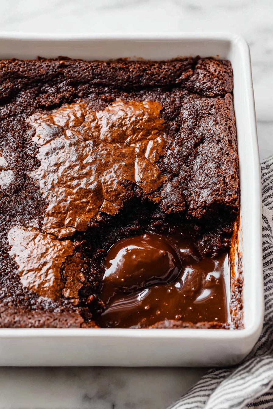 Decadent Hot Fudge Cake, hot fudge cake recipe, chocolate dessert, gooey chocolate cake, self-saucing pudding - A square white ceramic dish holds a rich chocolate dessert with a cracked, shiny top layer that is dark brown and slightly rough in texture. One section near the front corner is scooped out, revealing a thick, glossy, melted chocolate layer beneath the top crust. The melted chocolate is smooth and dark, contrasting with the more solid and matte top layer. The dish sits on a white marbled surface, and a striped cloth partially visible under one edge. photo taken with an iphone --ar 2:3 --v 7