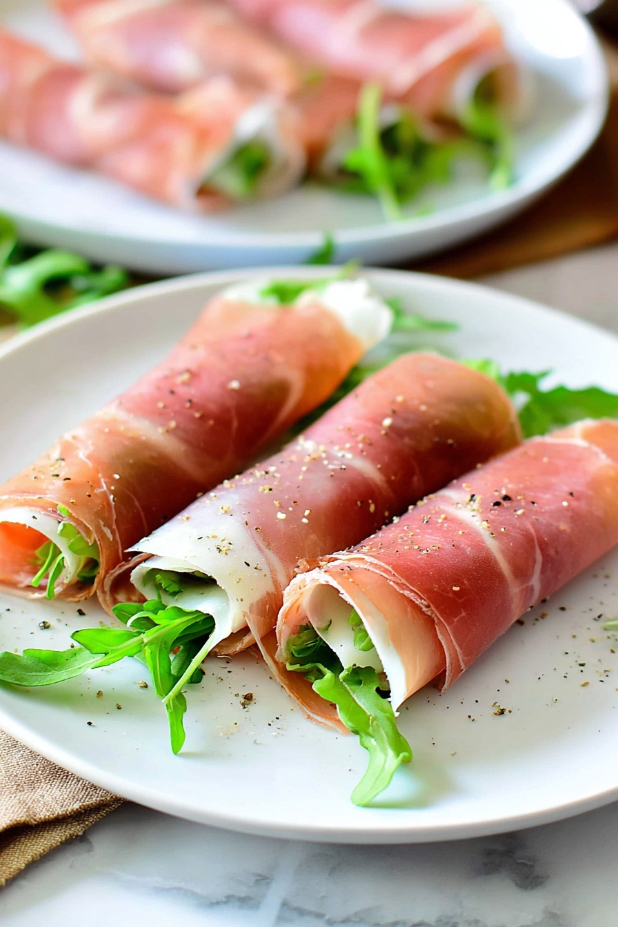 Fig and Prosciutto Roll-Ups, Fig and Prosciutto appetizer, easy fig prosciutto snacks, elegant appetizer ideas, prosciutto and goat cheese bites - Three thin rolled slices of pink prosciutto are placed on a white plate, each filled with green arugula leaves peeking out from one end. The prosciutto has a smooth, slightly shiny texture with light white fat streaks and is lightly sprinkled with coarse black pepper. The white plate sits on a white marbled surface with some leafy greens scattered around its edge. In the background, another white plate holds more similar prosciutto rolls, blurred to focus on the front plate. Photo taken with an iphone --ar 2:3 --v 7