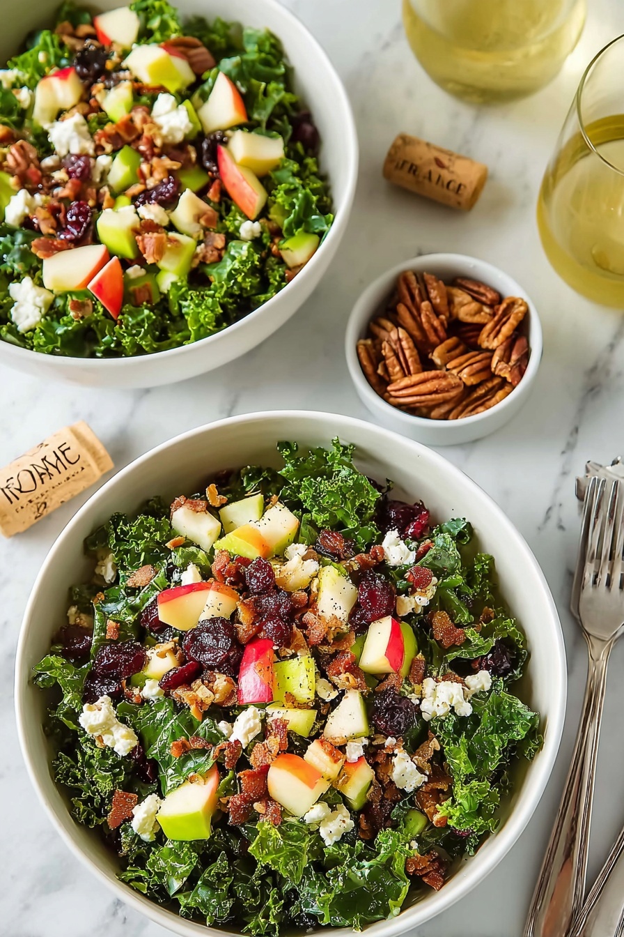 Apple Cranberry Pecan Salad with Candied Nuts, Apple Cranberry Pecan Salad, Holiday Fruit and Nut Salad, Fall Salad with Candied Pecans, Kale Fruit Nut Salad - Two white bowls filled with layered salad sit on a white marbled surface. Each bowl has a dense layer of dark green curly kale leaves at the bottom. On top of the kale are scattered pieces of red and yellow apple chunks with skin on, small light green celery bits, white crumbled cheese, dark red dried cranberries, and small brown pecan clusters. There are also small pieces of cooked bacon with a crispy texture mixed through the salad. Nearby, there is a small white bowl with extra pecans, a glass of light yellow drink, silver forks to the upper right, and a cork with the word 