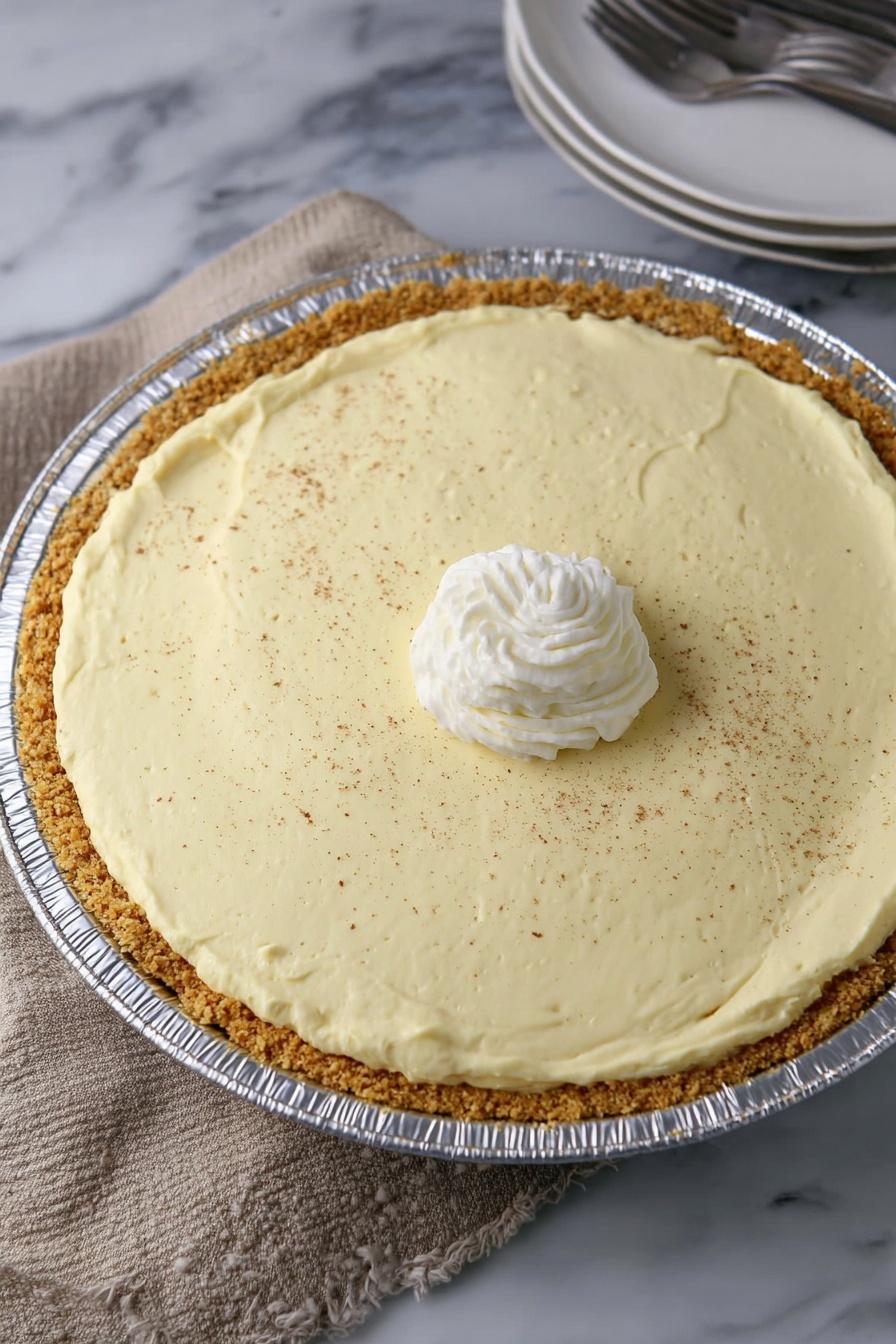 No-Bake Eggnog Pie, Eggnog Pie, Festive Holiday Desserts, Easy Holiday Pie, No-Bake Christmas Dessert - A creamy light yellow pie with one slice missing is in a shiny round aluminum pan. It has two layers: a thick light brown crumb crust at the bottom and a thick smooth creamy yellow filling on top. The pie is topped with a small swirl of white whipped cream in the center and sprinkled with fine brown powder all over. The pie sits on a light brown woven cloth with a white marbled surface showing at the edge. On the right side, a partial view of stacked white plates with a fork is visible. photo taken with an iphone --ar 2:3 --v 7