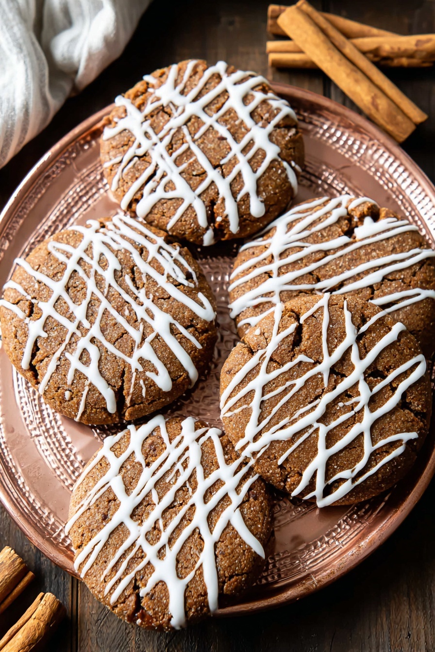 Soft Gingerbread Cookies, Gingerbread Cookies Recipe, Easy Gingerbread Cookies, Holiday Gingerbread Cookies, Chewy Gingerbread Cookies - A white plate holds eight round, light-brown cookies with a slightly cracked texture, each drizzled with thin white icing in a random, striped pattern across the tops. The cookies are evenly spaced on the plate, showing a soft and slightly raised surface with darker spots that hint at spices or chocolate bits. The plate sits on a white marbled surface, with a cinnamon stick and a pink cloth nearby, adding warm tones to the scene. Photo taken with an iphone --ar 2:3 --v 7