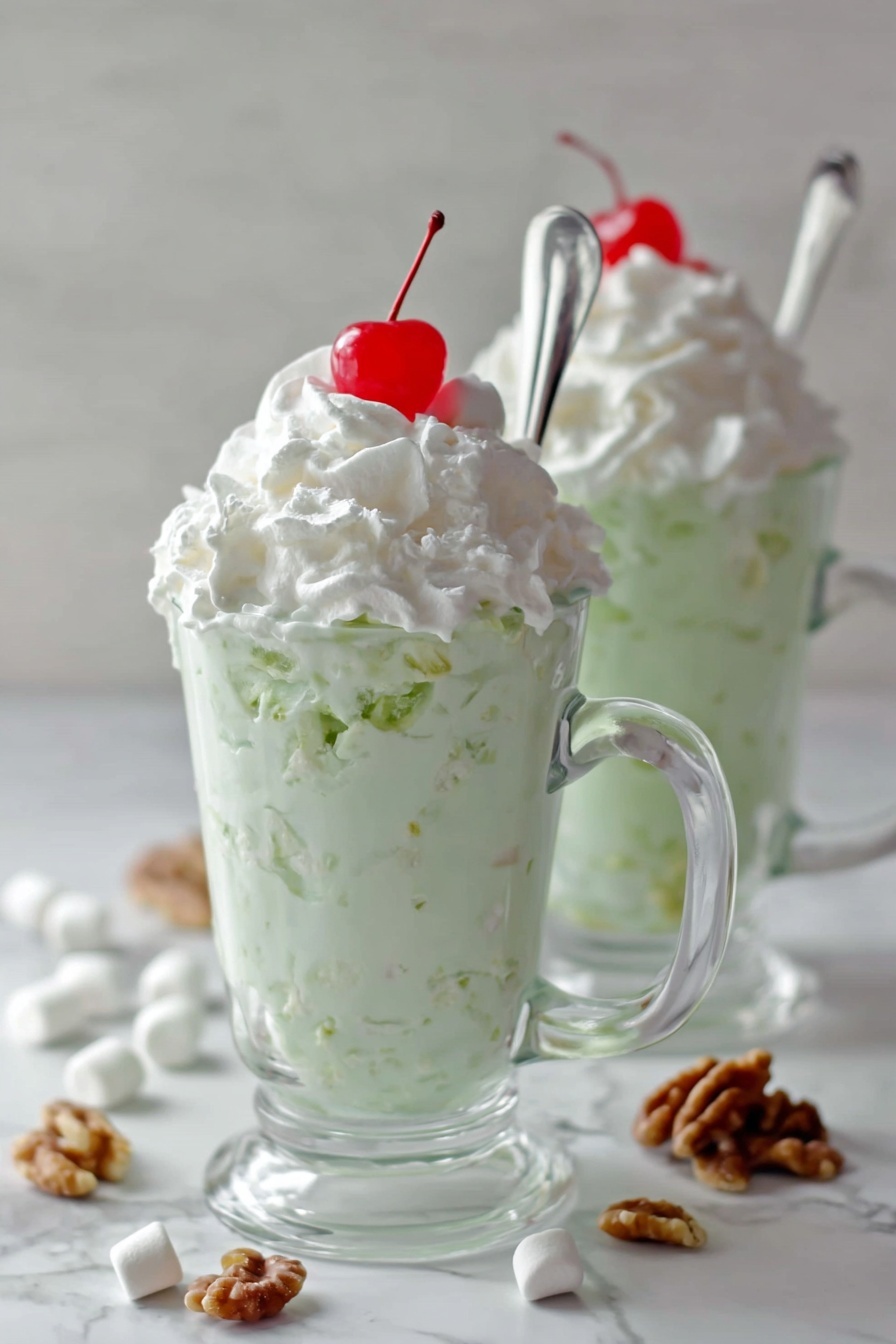 Easy Watergate Salad, Watergate Salad with Pineapple and Marshmallows, Pineapple Marshmallow Salad, Retro Watergate Salad, Simple Party Salad - Two clear glass mugs sit on a white marbled surface, each filled with a layered dessert. The bottom layer is a thick, creamy pale green mixture with visible small chunks, topped generously with a smooth white whipped cream layer. On the very top of each mound of whipped cream is a small, bright red cherry. Each mug has a long silver spoon inserted, leaning outwards. Around the mugs on the surface are scattered mini white marshmallows and small pieces of walnut. The photo taken with an iphone --ar 2:3 --v 7