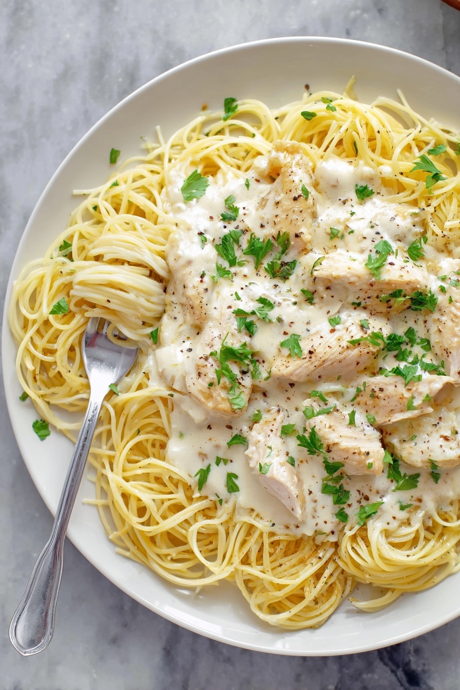 Creamy Crock Pot Angel Chicken, slow cooker chicken recipes, easy creamy chicken dinner, no-fuss crockpot chicken, beginner-friendly slow cooker meals - A white plate holds a dish of thin yellow spaghetti noodles arranged loosely in a circle. On top of the noodles are several pieces of light cream chicken, covered with a thick white creamy sauce that has small bits of herbs and black pepper sprinkled over it. Fresh green parsley leaves are scattered on top of the sauce for a touch of color. A silver fork rests on the right side of the plate, twirling some noodles with sauce. The plate sits on a white marbled surface. Photo taken with an iphone --ar 2:3 --v 7