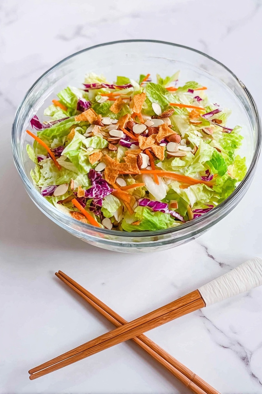 Crispy Asian Chicken Salad on Egg Roll Base, Asian chicken salad, crispy egg roll salad, quick Asian chicken recipe, Chinese-inspired chicken salad - A clear glass bowl sits on a white marbled surface, filled with a colorful salad made of several layers: the bottom layer has bright green lettuce leaves mixed with light green cabbage, scattered on top are thin orange carrot strips and bits of purple cabbage, then sprinkled with light brown almond slices and thin crispy golden strips, creating a fresh and crunchy look. Beside the bowl, a pair of wooden chopsticks with white tips rests on the surface. photo taken with an iphone --ar 2:3 --v 7