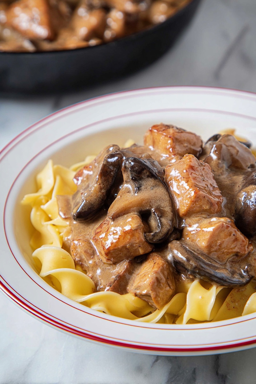 Easy Chicken Stroganoff, creamy chicken stroganoff, quick chicken dinner, comforting chicken recipes, family-friendly chicken dishes - The dish shows a layer of light yellow egg noodles at the bottom, with soft wide ribbons that curl slightly. On top, there is a thick layer of brown creamy sauce that coats chunks of cooked meat and slices of mushrooms. The meat pieces are uneven in size and have seared brown edges. The mushrooms are dark brown and smooth. Everything is served in a white bowl with a thin red ring around the edge. The bowl sits on a white marbled surface. In the background, there is a hint of a black skillet with more meat and sauce inside. Photo taken with an iphone --ar 2:3 --v 7