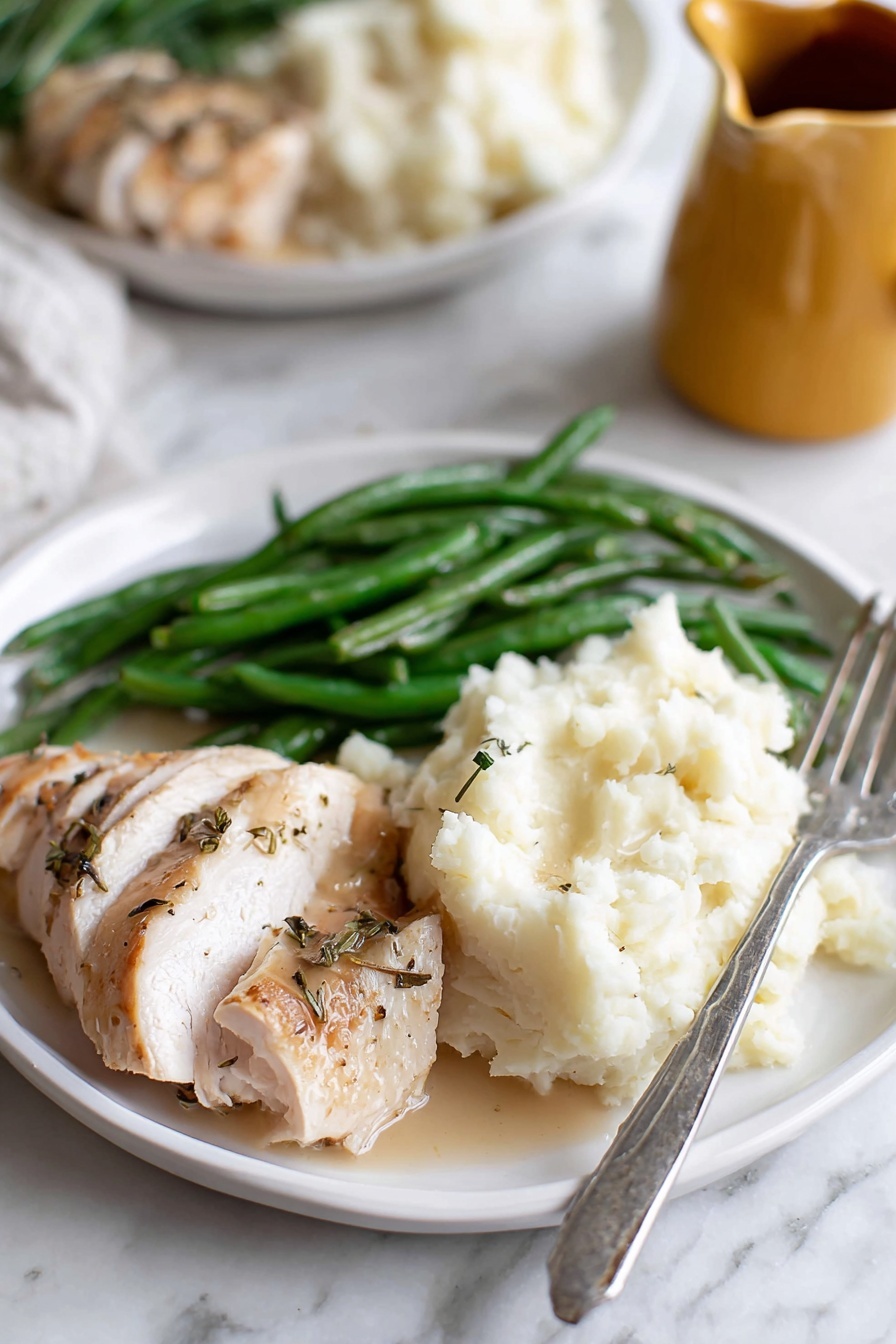 Homemade Turkey Gravy, turkey gravy, holiday gravy, easy turkey gravy, how to make turkey gravy - A white plate holds three main parts: on the left, green beans with a shiny, fresh texture; in the middle front, sliced cooked chicken with a light brown, slightly crispy skin and some herb seasoning; on the right, a large scoop of white mashed potatoes with a soft, fluffy texture, and a silver fork resting at the edge of the plate. The plate sits on a white marbled surface, with a blurred second plate in the back and part of a yellow jug visible. photo taken with an iphone --ar 2:3 --v 7