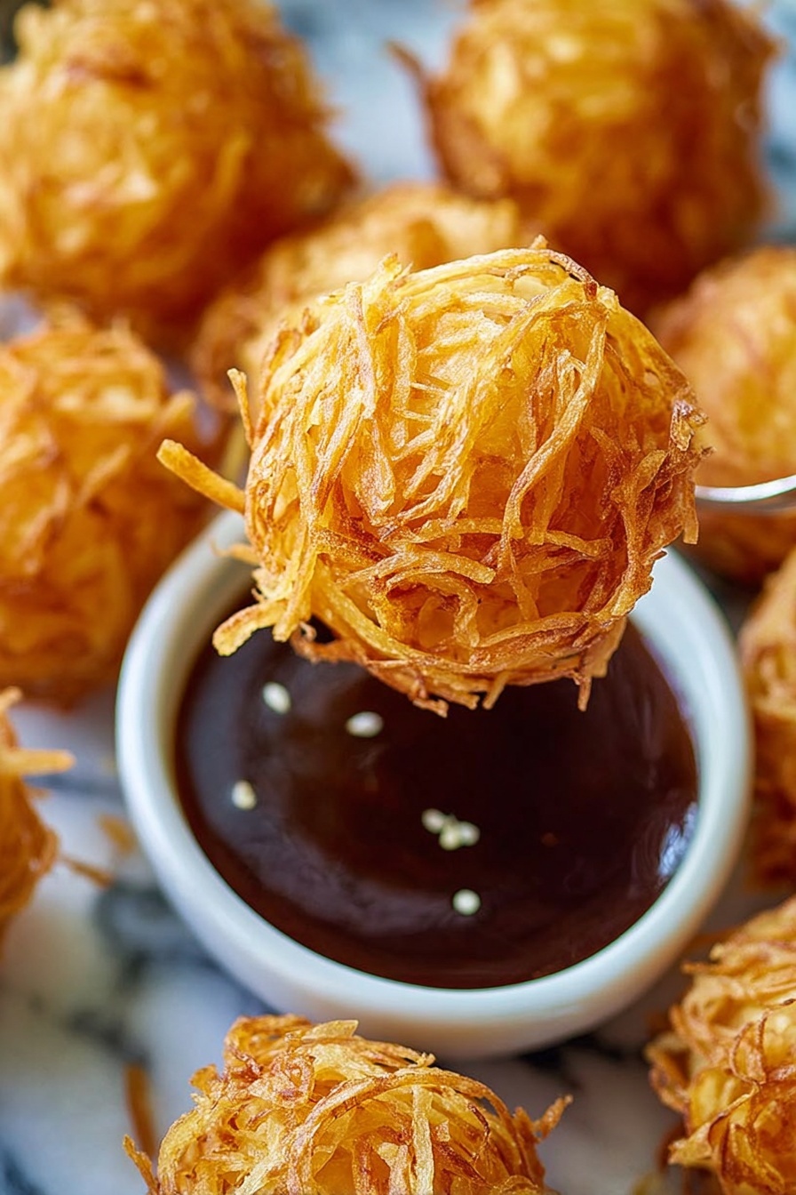 Crispy Fried Shrimp Balls, Shrimp Balls Recipe, Crispy Shrimp Appetizer, How to Make Shrimp Balls, Easy Shrimp Balls - The image shows several golden brown balls made from shredded, fried potato layers, giving them a textured and crispy look. One ball is held above a small white bowl filled with dark brown dipping sauce that appears glossy and smooth, with a few sesame seeds floating on top. The balls are arranged on a white marbled surface that adds a clean, bright background to the warm tones of the potato balls. The focus is sharp on the ball being held, showing the thin, curly potato strands wrapped around tightly, while the other balls and bowl fade softly into the background. photo taken with an iphone --ar 2:3 --v 7