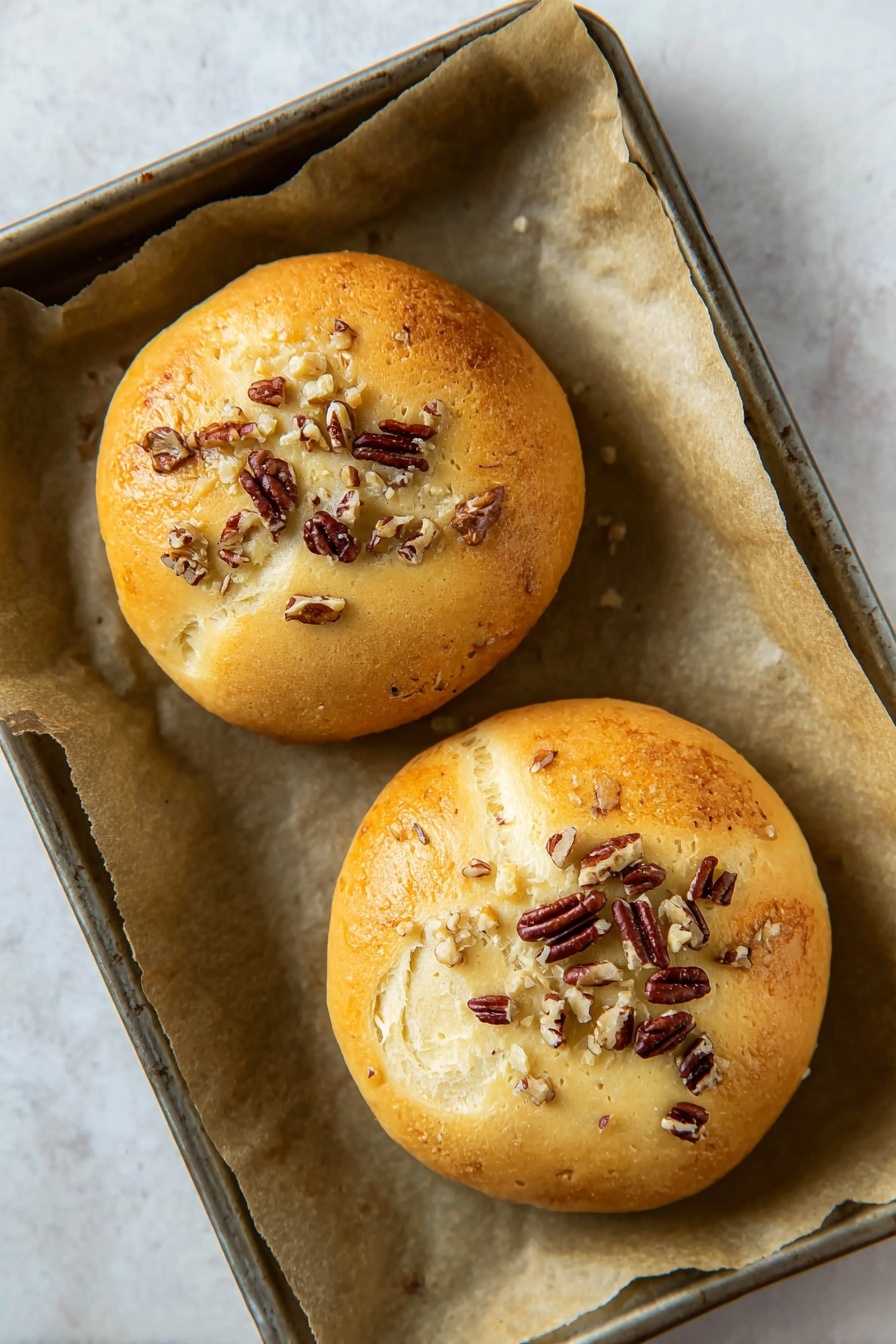 Cranberry Chicken Puffs, Cranberry Chicken Puffs recipe, easy cranberry chicken appetizer, savory cranberry chicken bites, quick crescent roll snacks - Two round golden brown buns rest on a baking tray lined with light brown parchment paper. Each bun has a slightly cracked top layer showing soft light beige dough beneath, sprinkled with chopped dark brown pecans scattered unevenly over the surface. The tray lies on a white marbled surface that is smooth and clean. Photo taken with an iphone --ar 2:3 --v 7