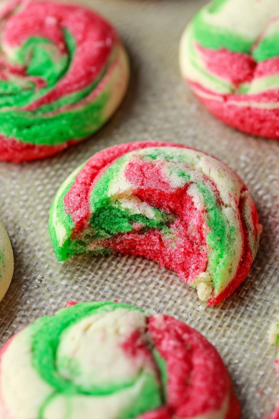 Peppermint Cheesecake Cookies, holiday peppermint cookies, festive cheesecake cookies, easy peppermint cookie recipe, Christmas dessert cookies - The image shows several round cookies with swirls of red, green, and white colors on a baking tray. One cookie is broken in half, showing its soft inside with the same bright red and green swirls mixed with white dough. The cookies have a slightly wrinkled texture and uneven edges, with the colors blending in a marbled pattern. The background is a white marbled texture photo taken with an iphone --ar 2:3 --v 7