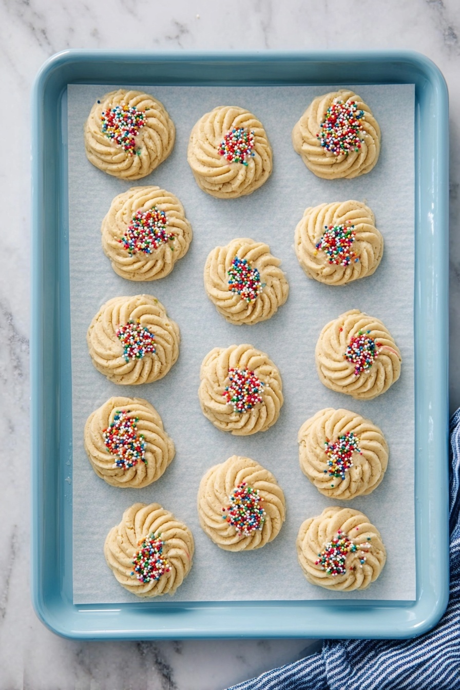 Best Butter Cookie, Butter Cookie Recipe, Homemade Butter Cookies, Classic Butter Cookies, Easy Butter Cookie Instructions - There are fifteen round cookies placed on parchment paper on a light blue baking tray. The cookies have a swirled pattern with textured ridges in a pale beige color. Nine of the cookies are sprinkled with colorful round sprinkles, while six cookies have no sprinkles and show only the smooth beige dough. The tray is set on a white marbled surface with a blue and white striped cloth partially visible on the right side. photo taken with an iphone --ar 2:3 --v 7