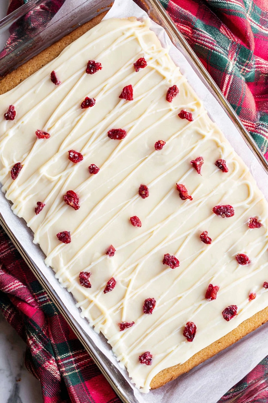 Cranberry White Chocolate Bliss Bars, cranberry white chocolate bars, holiday cranberry bars, easy cranberry dessert, white chocolate cranberry treats - A rectangular layered dessert lies on white parchment paper inside a glass baking dish on a white marbled surface. The bottom layer is golden brown, thin, and even. On top is a thick, smooth white frosting layer covering the entire surface. Thin white icing lines are drizzled across the frosting, forming a light grid pattern. Scattered over the top are small dried red fruit pieces, evenly spaced, adding bright red spots to the white frosting. The cloth nearby has green, red, and dark colors in a check pattern. photo taken with an iphone --ar 2:3 --v 7