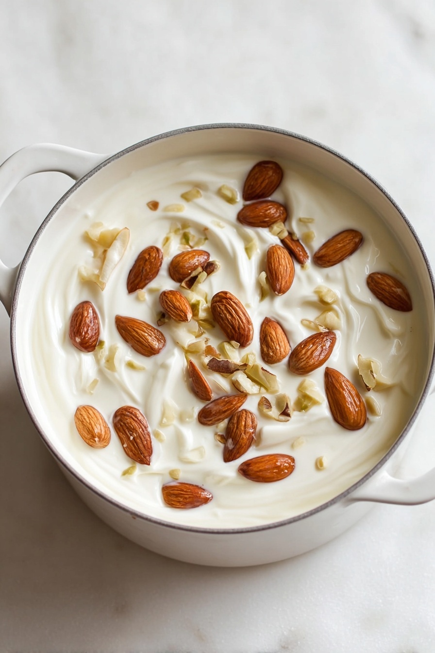 Homemade Italian Torrone Nougat, Italian Torrone Nougat, how to make torrone, traditional Italian nougat, homemade Italian candy - A white pot filled with one main layer of thick, creamy white yogurt mixed with whole and sliced almonds scattered evenly on the top. The almonds vary in shades of light brown to dark brown and are embedded slightly into the creamy yogurt, creating a textured surface with visible swirls in the yogurt beneath. The pot is sitting on a white marbled surface with soft lighting that highlights the smoothness of the yogurt and the natural shine of the nuts. Photo taken with an iphone --ar 2:3 --v 7