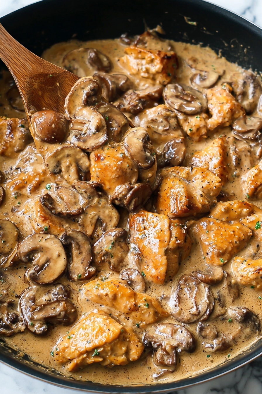 Easy Chicken Stroganoff, creamy chicken stroganoff, quick chicken dinner, comforting chicken recipes, family-friendly chicken dishes - The image shows a close-up of a pan filled with a creamy mushroom and chicken dish. The dish has two main layers: chunks of golden-brown chicken pieces and sliced brown mushrooms, both coated in a thick, light brown cream sauce. The chicken pieces are irregular in shape with a slightly crispy texture, while the mushrooms are soft and mixed throughout. A wooden spoon is partially submerged in the center, lifting some chicken and mushrooms. The pan is black and the background is a white marbled texture. Photo taken with an iphone --ar 2:3 --v 7