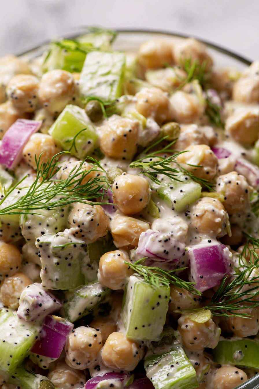 Healthy Tuna Chickpea Salad, Tuna Chickpea Salad, Healthy Salad Recipes, Quick Nutritious Meals, Protein-Packed Salads - The image shows a close-up of a salad with several layers and colors. The main elements are round chickpeas that are beige and smooth, mixed with small cubes of light green celery and purplish red onion. There are round, green capers scattered throughout. Fresh green dill sprigs are placed on top, adding a feathery texture. The salad is coated with a creamy, white dressing that has small black seeds mixed in. The entire dish sits against a white marbled surface in soft light. photo taken with an iphone --ar 2:3 --v 7