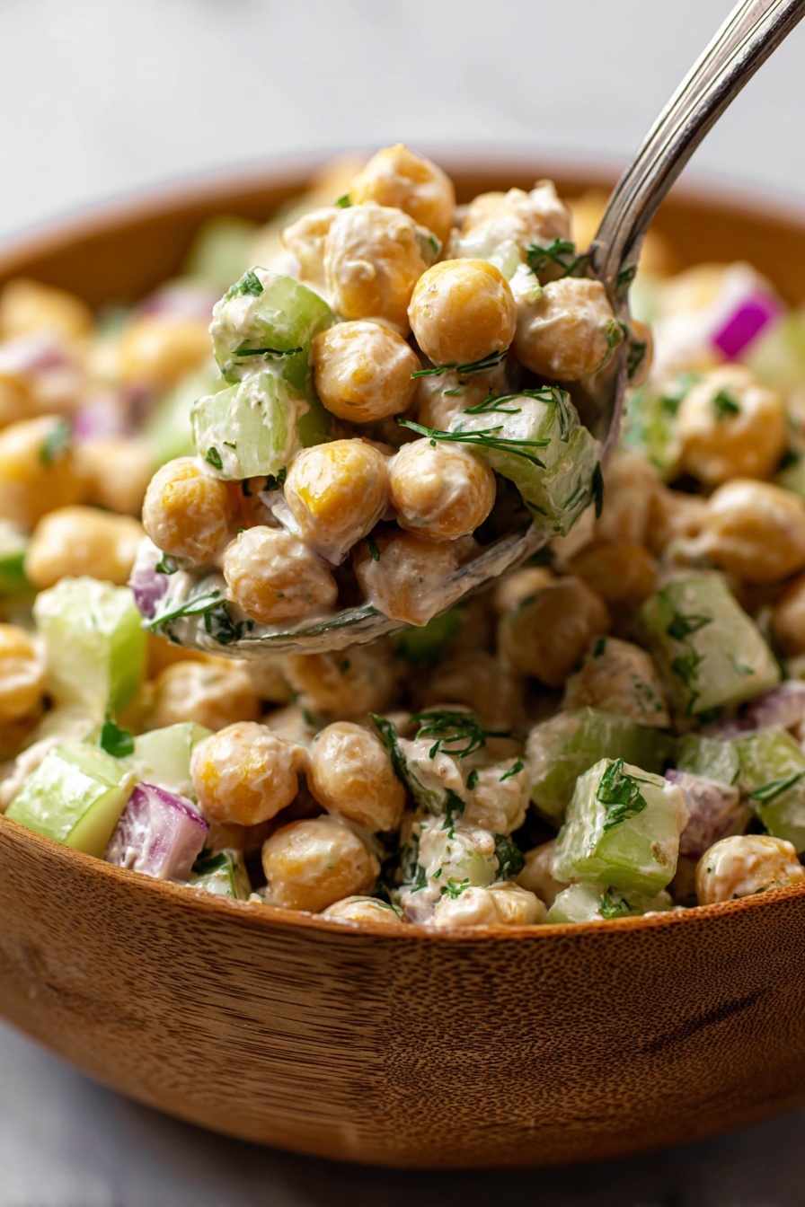 Healthy Tuna Chickpea Salad, Tuna Chickpea Salad, Healthy Salad Recipes, Quick Nutritious Meals, Protein-Packed Salads - The image shows a close-up of a chickpea salad served in a wooden bowl placed on a white marbled surface. The salad has three main layers: whole round chickpeas that are light golden and smooth, diced celery pieces that are pale green and crisp, and small bits of red onion adding a touch of purple. The ingredients are mixed with a creamy dressing that lightly coats everything, giving a soft texture. There are also small specks of fresh green herbs evenly spread throughout the salad. A metal spoon is lifting a portion of the salad from the bowl, with a woman's hand holding it, emphasizing the chunky and fresh look of the dish. photo taken with an iphone --ar 2:3 --v 7