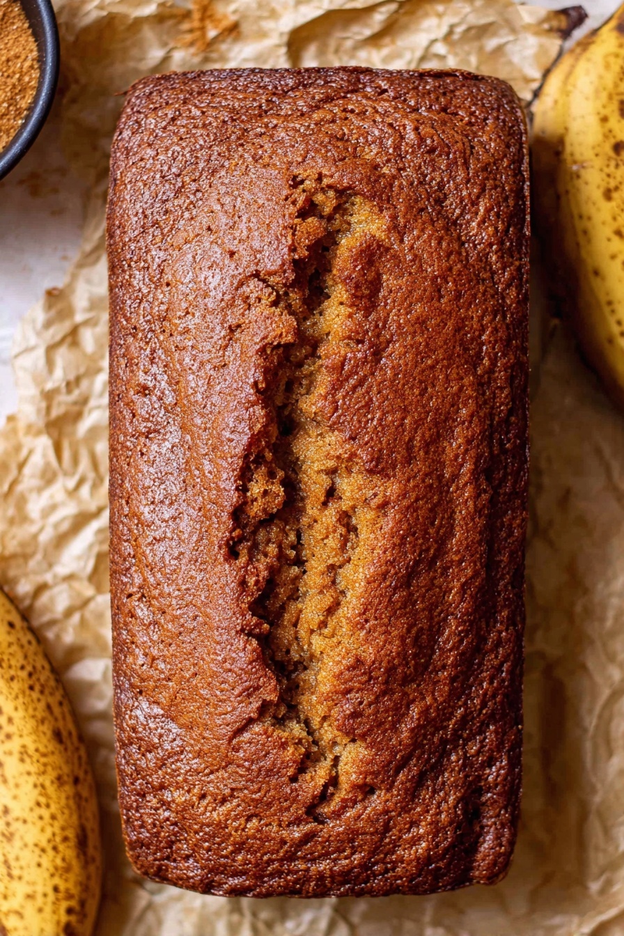 Moist Banana Bread, banana bread recipe, quick banana bread, easy banana bread, best banana bread - A single loaf of brown banana bread with a rough and cracked crust texture, sitting directly on crumpled parchment paper. The bread is rectangular with rounded corners and a deep crack running lengthwise down the middle. To the sides of the bread are parts of ripe bananas with dark spots, and part of a small black bowl with brown sugar is slightly visible on the left. The background is a white marbled texture. Photo taken with an iphone --ar 2:3 --v 7