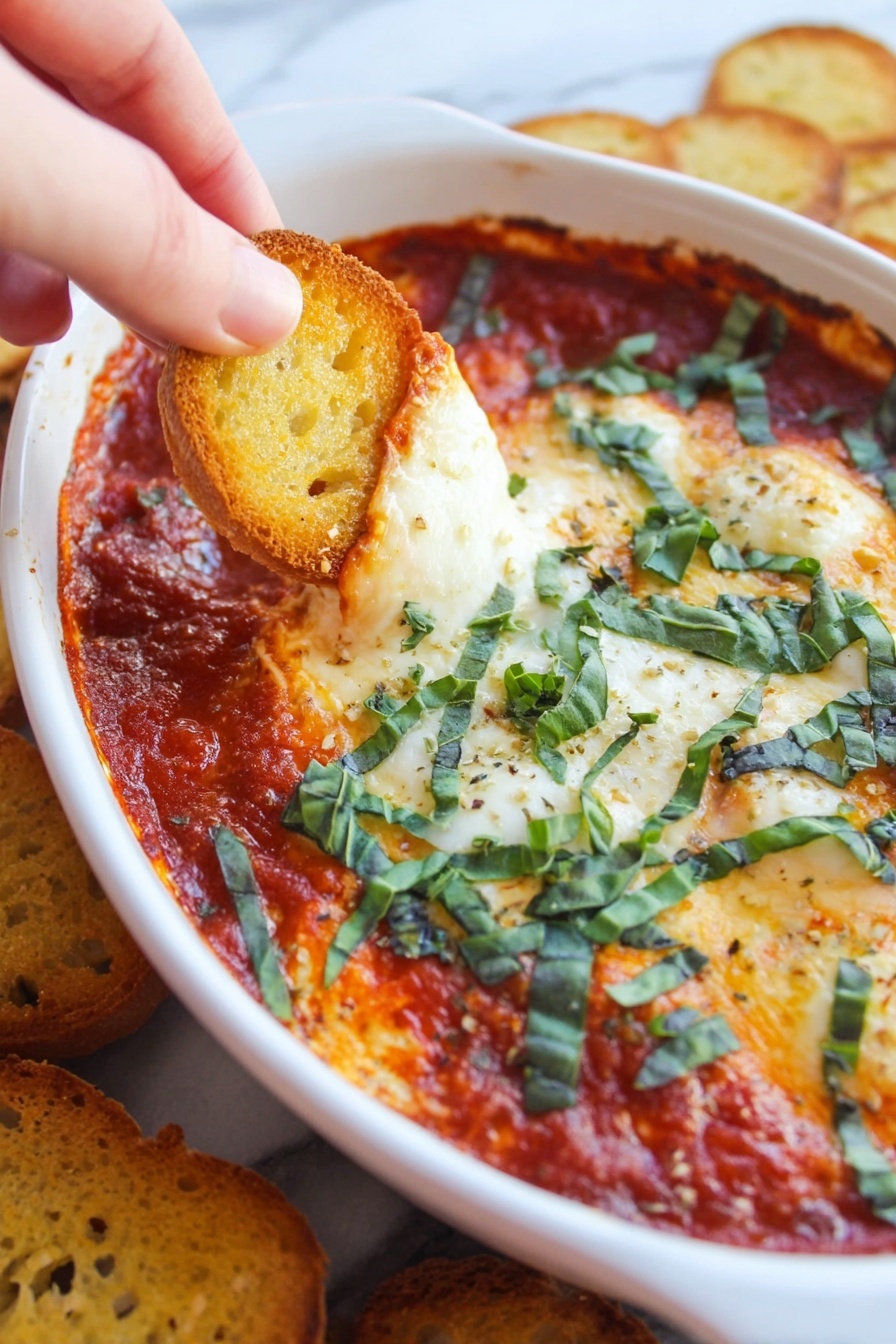 Goat Cheese Marinara Dip, easy appetizer with goat cheese and marinara, creamy goat cheese dip, quick savory dip recipes, crowd-pleasing party dip - A close-up image shows a white oval dish filled with a tomato sauce layer that is thick and red, topped with a baked layer of melted cheese that is golden and slightly browned. Thin strips of fresh green basil are scattered over the cheese and sauce, adding a fresh touch. A woman's hand is dipping a small, golden toasted bread slice into the sauce on the left side of the dish. The dish is placed on a white marbled surface with more toasted bread slices visible around it. photo taken with an iphone --ar 2:3 --v 7