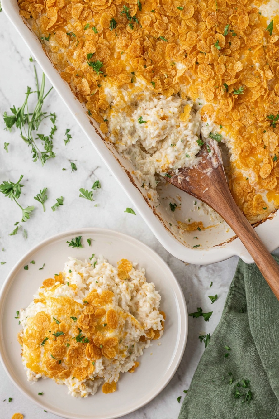 Cheesy Hashbrown Casserole, cheesy hashbrown casserole, cheesy potato casserole, hashbrown casserole recipe, cheesy breakfast casserole - A close-up view of a creamy casserole inside a white baking dish, showing two main layers: the bottom layer is a creamy white rice mixture with a soft texture, and the top layer is a golden, crunchy cornflake topping with a few small green herb pieces sprinkled on it. A wooden spoon inside the dish scoops out a portion, revealing the soft rice underneath the crunchy top. Next to the dish, a white plate holds a serving of the casserole, displaying the same two layers with the crunchy cornflakes on top and creamy rice underneath, also garnished with small green herbs. The whole scene sits on a white marbled surface with scattered green herb leaves and a green cloth napkin on the side. Photo taken with an iphone --ar 2:3 --v 7
