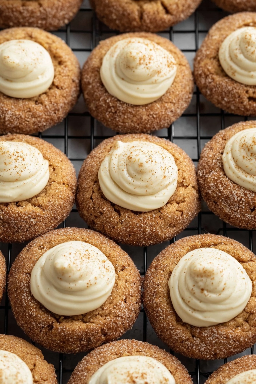 Eggnog Snickerdoodle Thumbprint Cookies, holiday eggnog cookies, cinnamon sugar thumbprint cookies, festive cookie recipes, creamy eggnog filling cookies - The image shows many round cookies arranged closely on a black cooling rack, placed against a white marbled background. Each cookie has two layers: the base is a golden brown cookie with a rough texture covered in a light dusting of sugar and cinnamon, and on top, there is a smooth, creamy white dollop of frosting in the center of each cookie. The frosting is thick with a swirl pattern and has small specks of cinnamon sprinkled over it, giving a contrast between the crispy cookie and soft frosting. photo taken with an iphone --ar 2:3 --v 7
