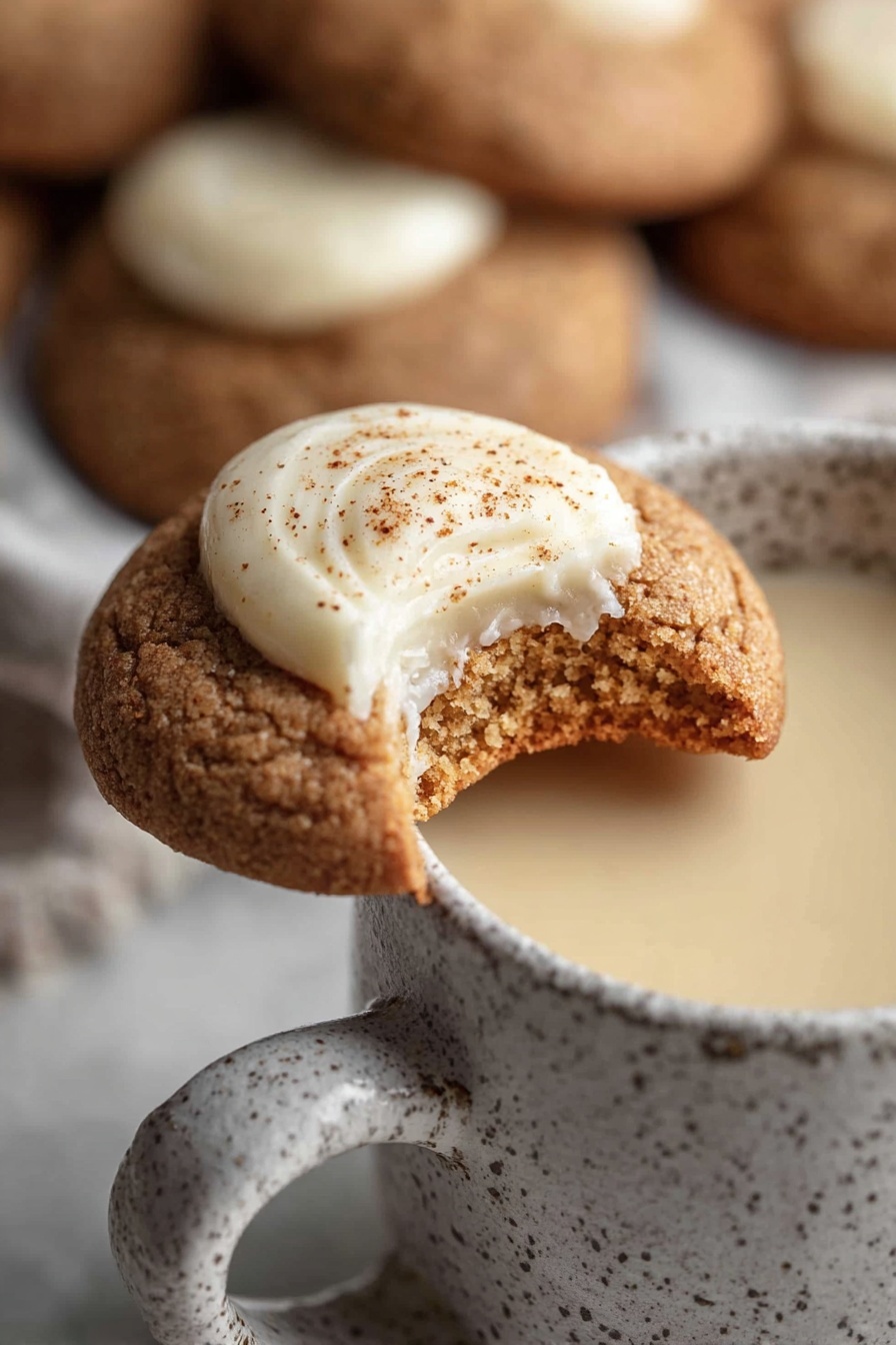 Eggnog Snickerdoodle Thumbprint Cookies, holiday eggnog cookies, cinnamon sugar thumbprint cookies, festive cookie recipes, creamy eggnog filling cookies - A close-up image shows a light brown cookie with a rough texture, topped with a dollop of smooth, creamy white frosting sprinkled with fine brown specks. The cookie has a visible bite taken from its right side, revealing a dense inner crumb. It rests on the rim of a white speckled mug filled with a creamy beige liquid. The background features several similar cookies blurred out, placed over a white marbled surface. photo taken with an iphone --ar 2:3 --v 7