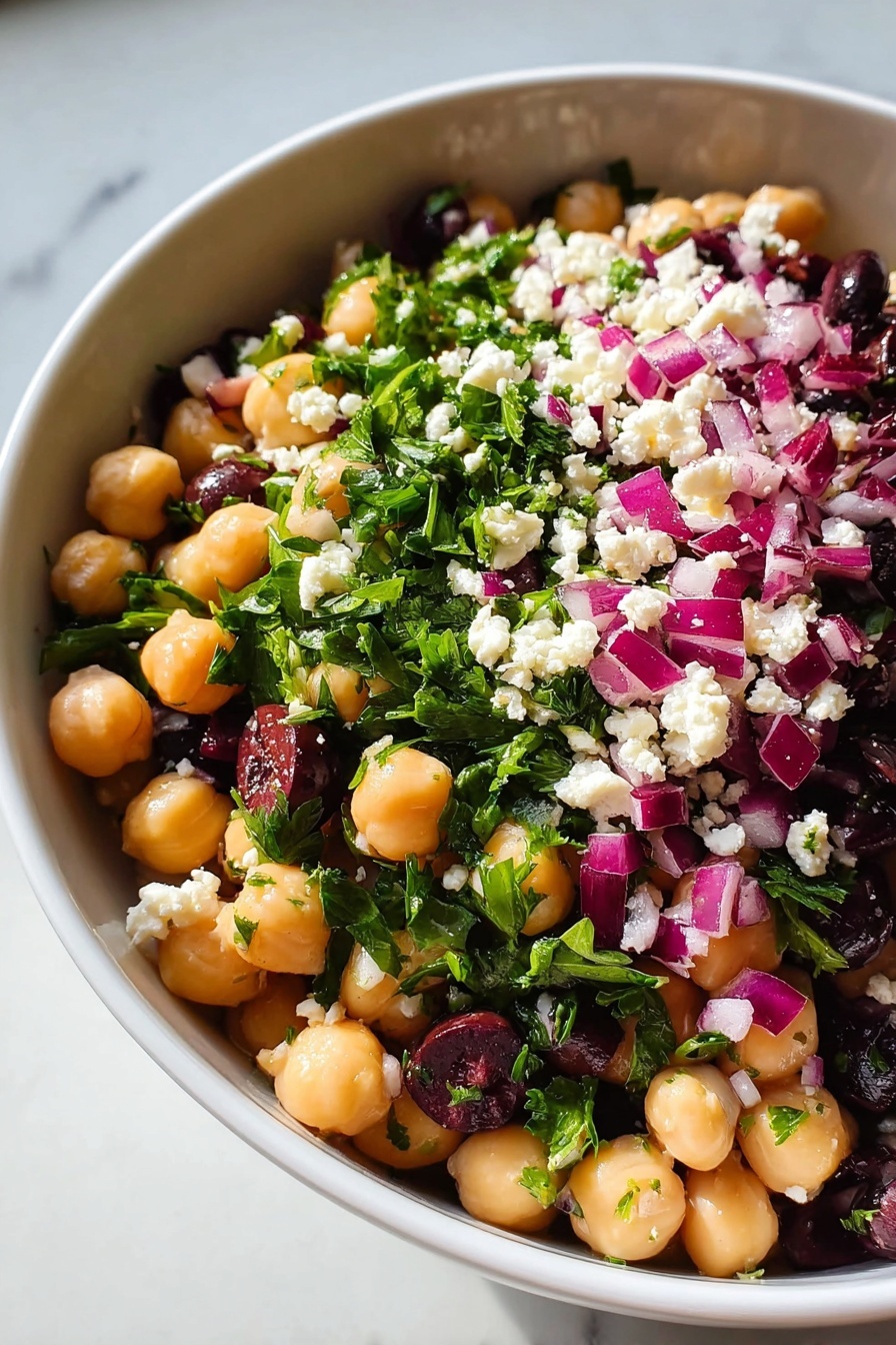 Chickpea Bean Salad with Feta, healthy chickpea salad, easy feta bean salad, vegetarian bean salad recipe, Mediterranean chickpea salad - A close-up view of a white bowl filled with a colorful chickpea salad, showing roughly three layers. The bottom layer consists of smooth, round pale beige chickpeas mixed with dark purple olives. The middle layer has bright green chopped leaves, likely parsley, scattered throughout. The top layer is dotted with small white cheese crumbles and chopped purple-red onions, adding texture and vibrant color contrast. The bowl sits on a white marbled surface. photo taken with an iphone --ar 2:3 --v 7