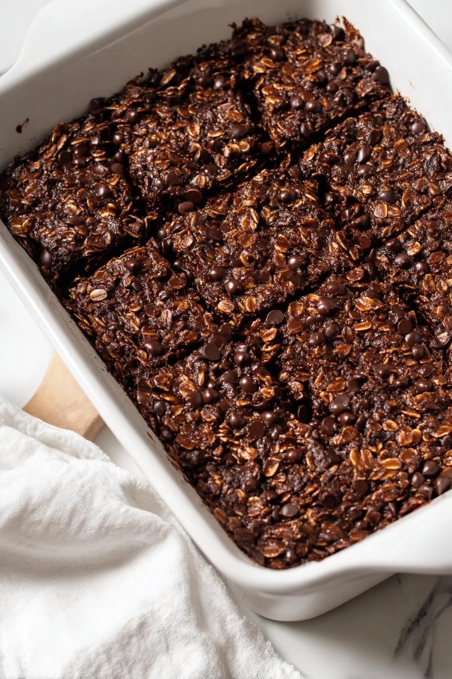 Chocolate Banana Baked Oatmeal, healthy baked oatmeal recipes, easy breakfast ideas with bananas, chocolate breakfast dishes, wholesome breakfast recipes - A rectangular white ceramic dish filled with a baked dark brown oat and chocolate chip mixture that is thick and textured. The surface is rough with visible oats and small shiny chocolate chips scattered throughout. The dish is placed on a white marbled surface with a white cloth partially visible at the bottom left. The top is divided into six uneven square pieces that show the dense and moist texture inside. Photo taken with an iphone --ar 2:3 --v 7