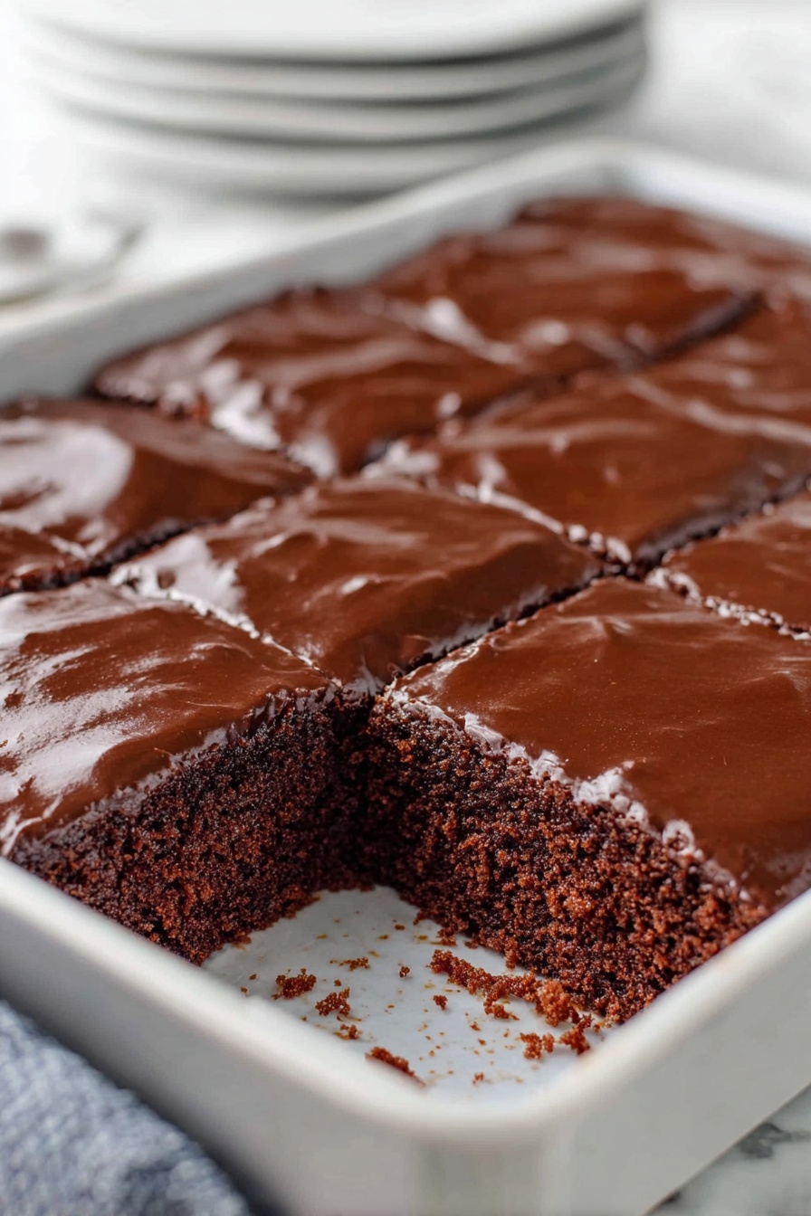 Texas Sheet Cake with Chocolate Frosting, moist chocolate cake recipe, easy Texas sheet cake, quick chocolate sheet cake, crowd-pleasing chocolate cake - A close-up of a chocolate sheet cake with two layers, the bottom layer is a soft, dark brown cake with a slightly crumbly texture, and the top layer is a shiny, smooth chocolate glaze covering the entire surface. The cake is cut into squares, and one piece is missing, revealing the inside texture. The cake is inside a white baking pan set on a white marbled surface, with parts of a woman's hand visible holding the pan. In the background, there is a soft blur of stacked white plates. photo taken with an iphone --ar 2:3 --v 7