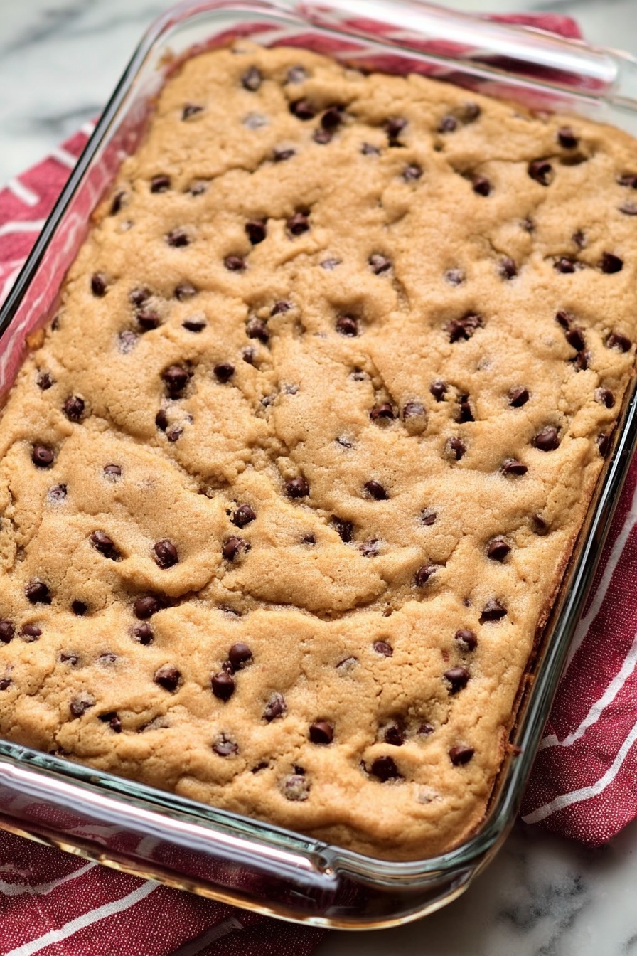 Caramel Chocolate Chip Bars, caramel chocolate chip bars, chocolate caramel bars, easy caramel dessert, chewy chocolate bars - A glass rectangular baking dish filled with one thick layer of golden brown, slightly uneven baked cookie dough, dotted with small dark chocolate chips. The cookie surface shows a soft, bubbly texture with some parts more raised and others flat, creating an inviting mix of crispy and soft areas. The dish sits on a white marbled surface with a red and white striped cloth peeking from underneath one side. Photo taken with an iphone --ar 2:3 --v 7