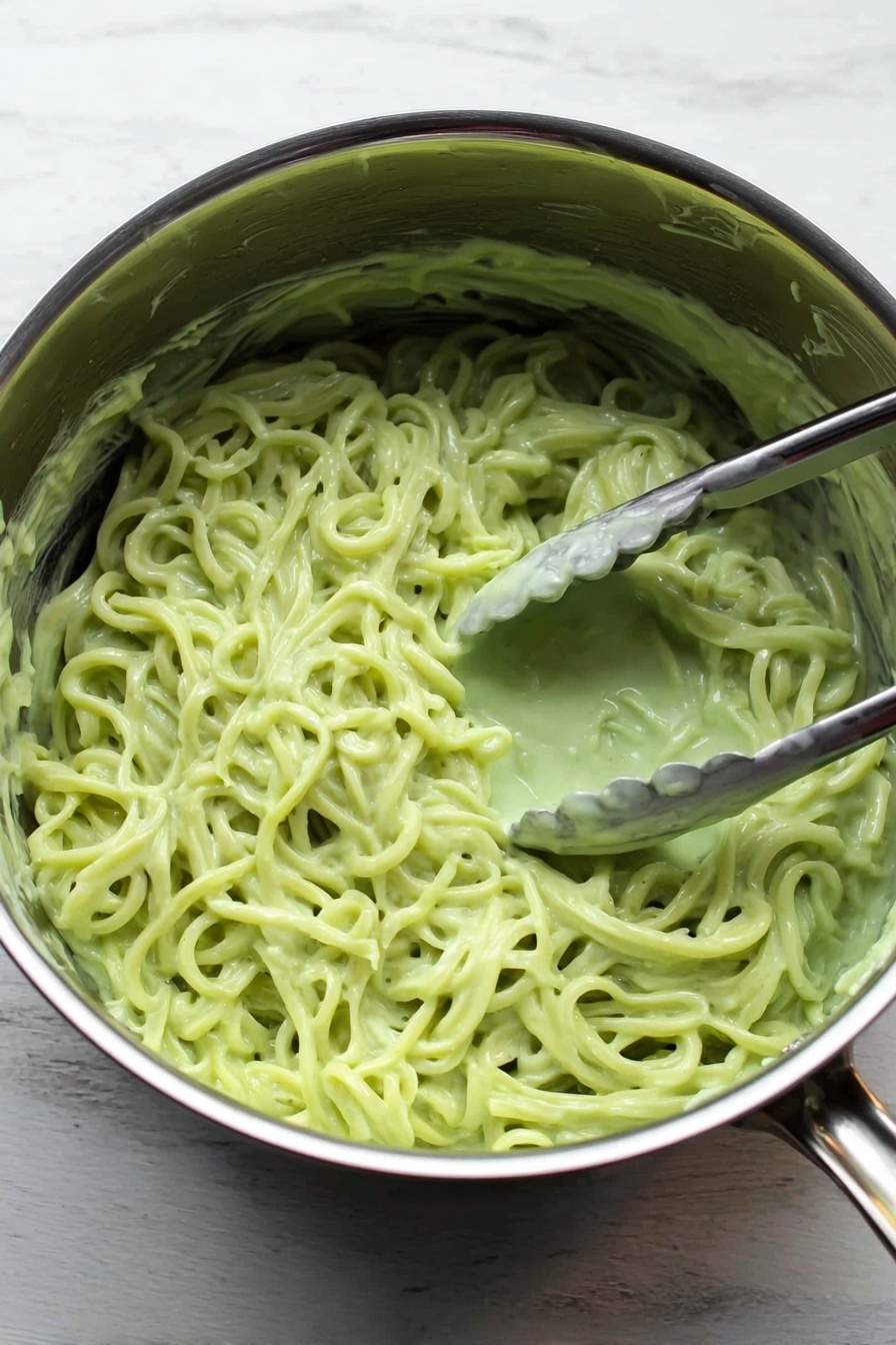Vegan Cilantro Lime Noodles, vegan noodle recipes, healthy plant-based noodles, quick vegan dinners, cilantro lime pasta - Inside a shiny silver pot placed on a white marbled surface, there is a mass of noodles covered in a thick, light green sauce. The sauce is creamy and clings to the noodles, giving them a smooth texture. A silver tong is partially dipped into the noodles on the right side of the pot, with a bit of sauce stuck on the tong. The noodles have a slightly twisted shape and fill most of the pot inside. Photo taken with an iphone --ar 2:3 --v 7