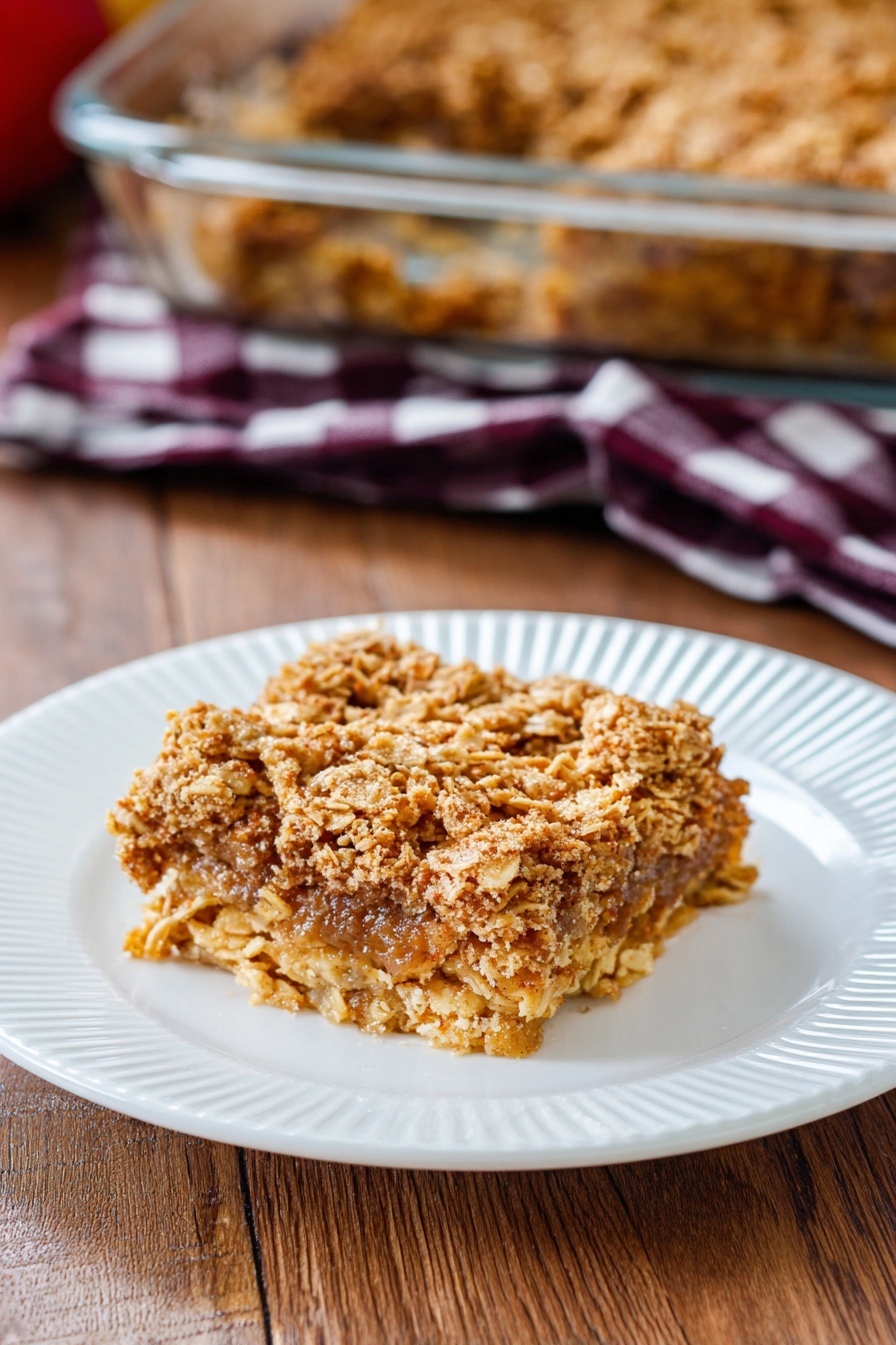 Apple Cinnamon Baked Oatmeal, healthy baked oatmeal breakfast, easy apple cinnamon brunch, cozy breakfast recipes, wholesome apple baked oats - A single square piece of oat crumble dessert sits in the center of a white plate with ridged edges. The dessert has two main layers: the top layer is golden brown with a rough texture made of baked oats mixed with cinnamon, while the bottom layer appears softer and darker, showing a moist filling beneath the oats. The plate is placed on a wooden table, and in the background, there is a glass baking dish with more of the same oat crumble. A dark red and white checkered cloth is also slightly visible behind the plate. Photo taken with an iphone --ar 2:3 --v 7