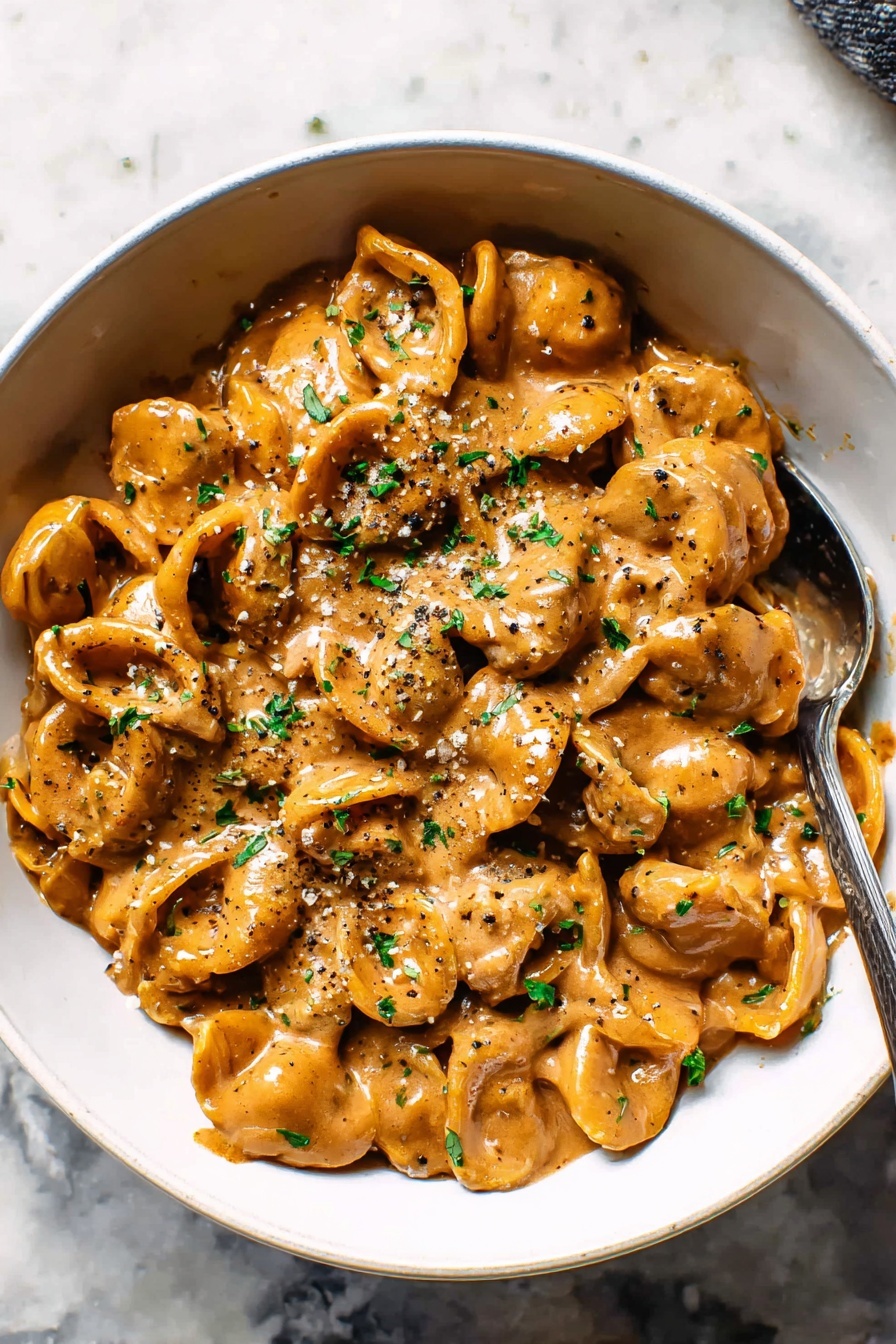 French Onion Pasta with Melted Gruyere, French Onion Pasta, cheesy pasta with caramelized onions, creamy Gruyere pasta, French-inspired pasta dish - The image shows a white bowl filled with small, shell-shaped pasta coated in a thick, creamy brown sauce. The pasta appears soft and layered evenly in the bowl with specks of black pepper and chopped green herbs sprinkled on top for contrast. A silver spoon is partially dipped into the pasta toward the right side of the bowl. The bowl sits on a white marbled surface with soft natural lighting highlighting the shiny texture of the sauce photo taken with an iphone --ar 2:3 --v 7