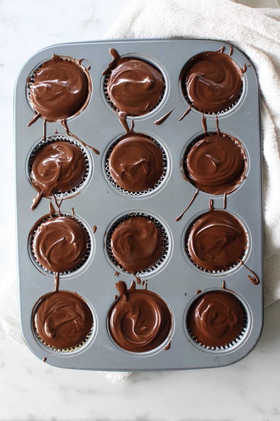 No Bake Peanut Butter Oat Cups, peanut butter oat treats, no bake snack recipes, chocolate peanut butter cups, easy healthy snacks - A gray muffin tray with twelve round cups, each filled with a smooth, glossy layer of melted chocolate that slightly spills over the edges. The chocolate has a shiny, swirled texture on top, and thin chocolate drips and streaks connect some of the cups and run down the tray’s sides. The tray rests on a white marbled surface with a soft white cloth partially visible at the top right corner. Photo taken with an iphone --ar 2:3 --v 7