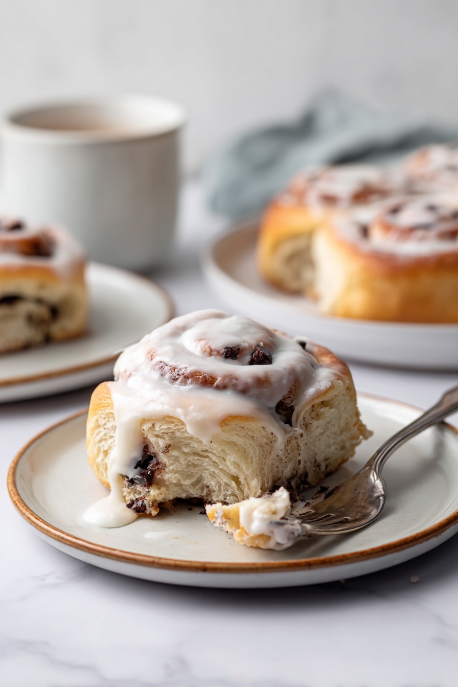 S’mores Rolls with Toasted Meringue, s'mores dessert, homemade s'mores, toasted meringue recipe, gooey chocolate rolls - The image shows a single cinnamon roll with visible layers of soft dough filled with dark chocolate chunks, placed on a white plate with a light brown rim. The top layer of the cinnamon roll is covered with white icing that drips down the sides, creating a creamy texture. Part of the roll is pulled away by a fork lying next to it, revealing the inner swirls with chocolate pieces. In the background, more cinnamon rolls on white plates and a white cup are softly blurred, all set against a white marbled surface. Photo taken with an iphone --ar 2:3 --v 7