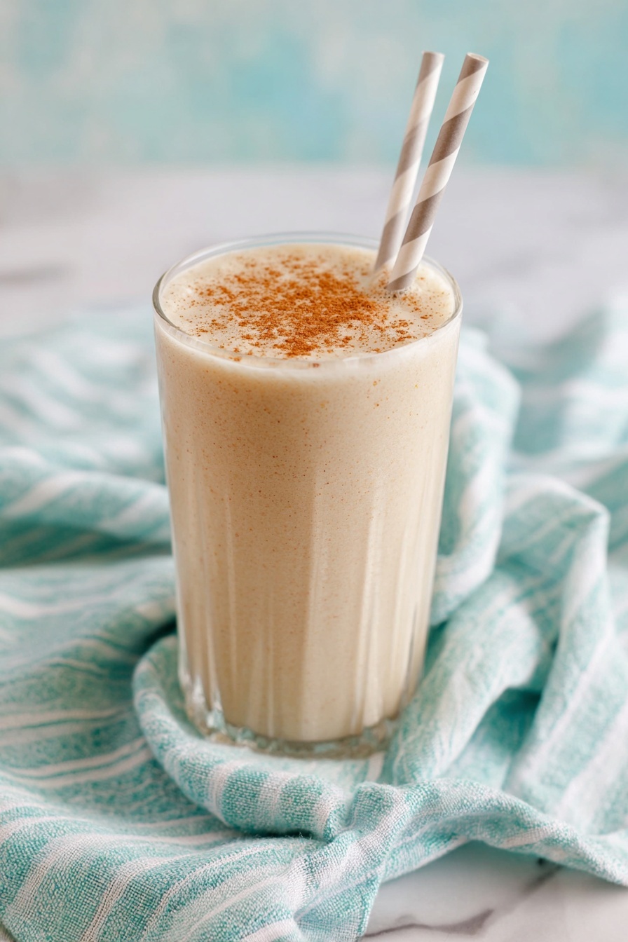 Vanilla Chai Protein Shake, healthy chai protein smoothie, quick breakfast protein shake, energizing chai latte protein, cozy vanilla chai smoothie - A tall clear glass filled with a creamy, light beige drink that has small specks mixed throughout, showing a smooth and thick texture. The top of the drink is sprinkled evenly with a light brown powder. The glass sits on a green and white striped cloth on a white marbled surface. The background is soft and out of focus with light creamy tones. photo taken with an iphone --ar 2:3 --v 7
