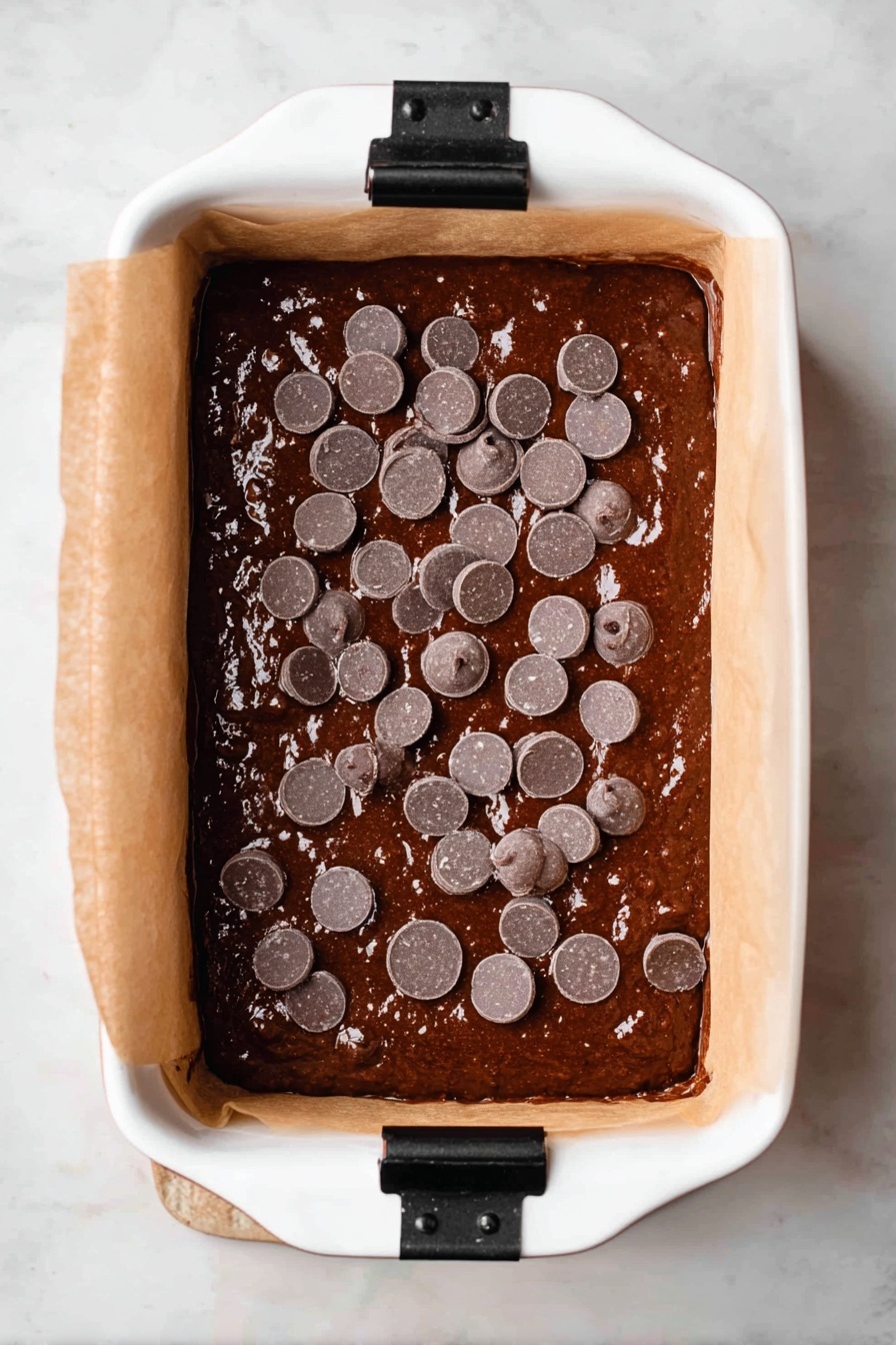 Chocolate Espresso Banana Bread, banana bread with espresso and chocolate, easy chocolate banana bread, moist banana bread with coffee, homemade chocolate banana loaf - The image shows a white rectangular baking dish lined with parchment paper, filled with thick dark brown chocolate batter. On top of the batter, there are many round, flat, shiny chocolate chips scattered unevenly. The parchment paper extends up on the longer sides, held in place with black clips. The dish is placed on a white marbled surface that contrasts with the rich color of the batter. The texture of the batter looks smooth and glossy, with small air bubbles visible. photo taken with an iphone --ar 2:3 --v 7