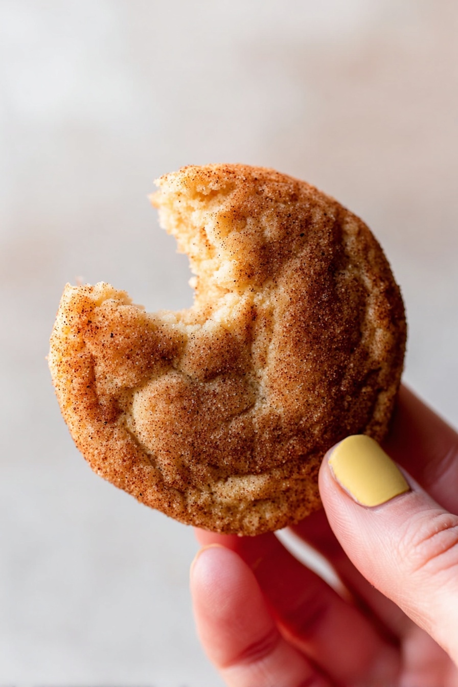 Brown Butter Snickerdoodle Cookies, gluten-free snickerdoodles, soft chewy cookie recipes, easy cookie recipes with browned butter, classic cinnamon sugar cookies - A close-up shows a woman's hand holding a round cinnamon sugar cookie with a bite taken out of the top. The cookie has a golden-brown color with a visible coating of cinnamon sugar that gives a slightly rough texture. The woman's thumb has a yellow nail polish, and the plain white marbled background softly blurs behind the cookie, drawing attention to the details on the cookie's surface and the soft texture where it was bitten photo taken with an iphone --ar 2:3 --v 7