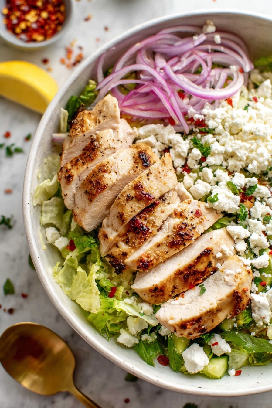 Greek Chicken Bowls with Tzatziki, Greek chicken bowls, Mediterranean chicken bowls, healthy Greek lunch, easy Greek dinner - The dish shows a white bowl filled with several layers starting with a bed of finely chopped green lettuce at the bottom. On top of the lettuce, there are thin slices of purple onion scattered along with some red pepper flakes and small pieces of fresh green herbs. The main layer is made of evenly sliced cooked chicken pieces, lightly browned on the edges and laid neatly in a row across the bowl. Crumbled white cheese is sprinkled generously all over the chicken and vegetables. A lemon wedge sits on the edge near the lettuce. The bowl is placed on a white marbled surface with some scattered red pepper flakes and herbs around it. A gold spoon rests inside the bowl near the chicken. photo taken with an iphone --ar 2:3 --v 7